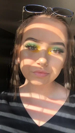 Woman with colorful eye makeup and sunglasses on head, wearing striped top, indoor selfie.