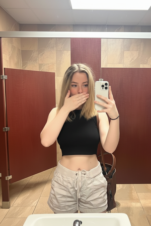 Woman in black crop top and beige shorts takes mirror selfie in bathroom.