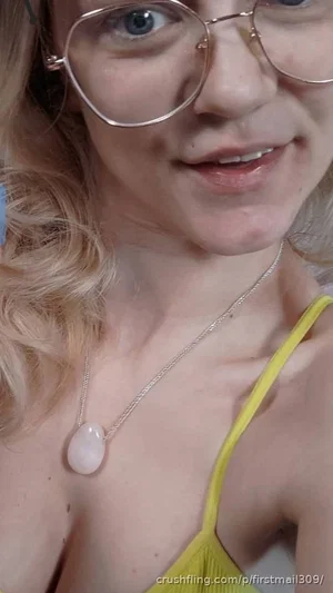 Woman with blonde hair and glasses wearing a yellow top and necklace, close-up selfie.