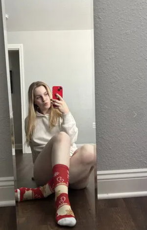 Blonde woman in white hoodie and red socks taking mirror selfie indoors.