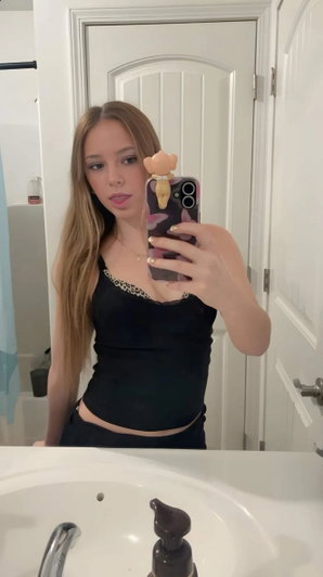 Blonde woman taking mirror selfie in bathroom wearing black top.