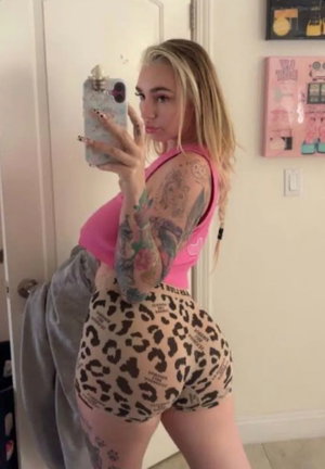 Blonde woman in pink top and leopard shorts taking mirror selfie indoors.