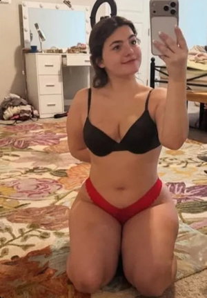 Curvy woman in black bra and red panties taking selfie in bedroom.