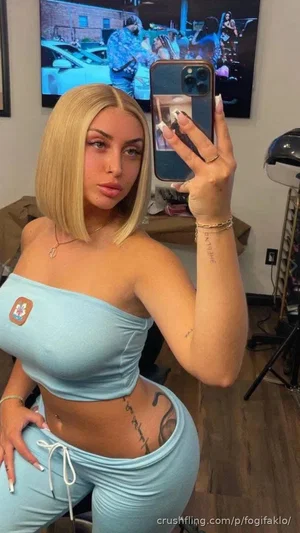 Woman with blonde bob, light blue crop top and matching pants, taking mirror selfie indoors.