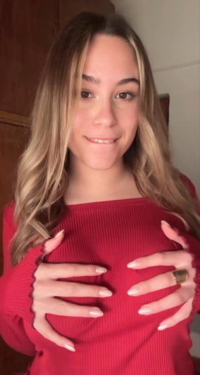 Woman with long blonde hair in red off-shoulder top, hands on chest, smiling indoors.
