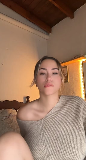 Woman in off-shoulder sweater sitting indoors, looking at camera.