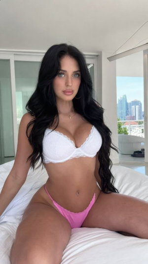Woman with long dark hair in white bra and pink underwear sitting on bed indoors.