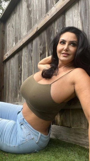 Woman with dark hair in olive crop top and jeans, leaning against wooden fence outdoors.