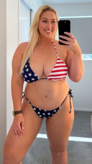 Woman with blonde hair in American flag bikini taking mirror selfie indoors.