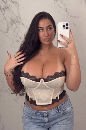 Woman with long dark hair in lace corset and jeans, posing in front of marble wall.