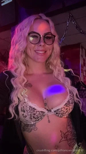 Woman with blonde curly hair, glasses, tattoos, wearing lace bra and black jacket in dimly lit