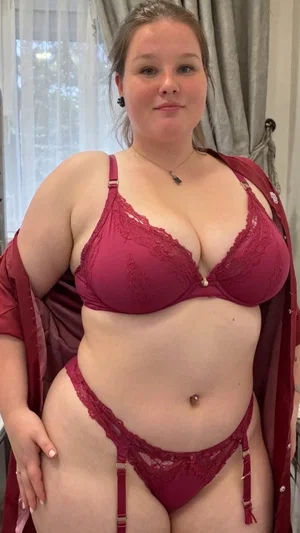 Woman in maroon lace lingerie and robe, standing indoors near window.