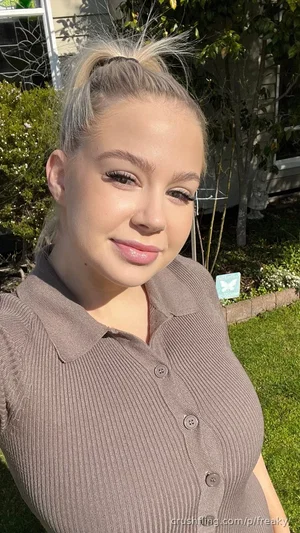 Woman with blonde hair in a ponytail, wearing a brown ribbed button-up top, outdoors in a garden.