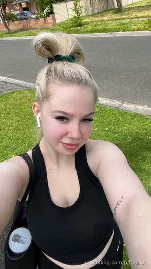 Woman with blonde hair in bun, wearing black tank top, standing outdoors on grass near street.
