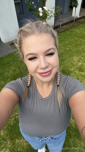 Woman with blonde braids in gray t-shirt and jeans, smiling in a grassy outdoor setting.