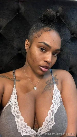 Woman with tattoos wearing gray lace-trimmed top, sitting against black tufted headboard.