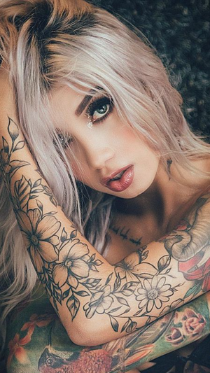 Tattooed blonde woman with makeup posing on dark surface.
