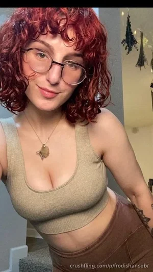 Woman with red curly hair and glasses wearing a beige crop top and brown pants indoors.
