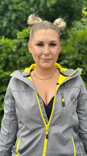 Woman with blonde hair in buns, wearing gray and yellow zip-up jacket over black top, outdoors with