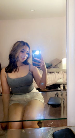 Curvy woman taking mirror selfie in bedroom wearing tank top and shorts.