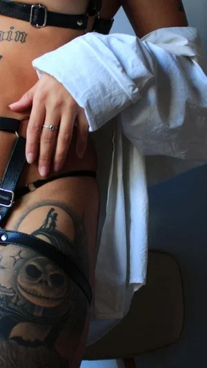 Woman with tattoos wearing black harness and white shirt, hand on hip.