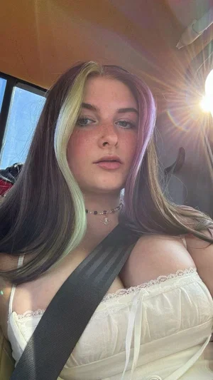 Woman with colorful hair in white lace top, sitting in car with seatbelt on.