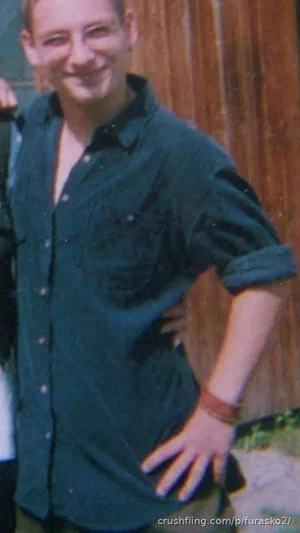 Man in dark button-up shirt, smiling, standing outdoors near wooden wall.