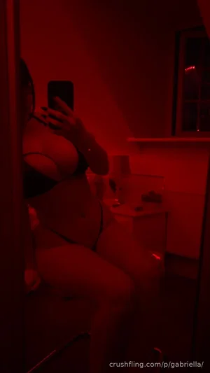 Woman in black lingerie taking mirror selfie in dim red-lit room.