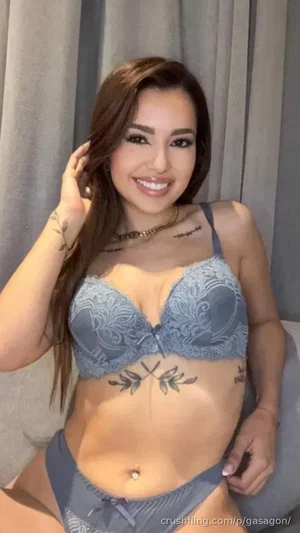 Woman in gray lace lingerie posing indoors with tattoos visible on torso and arm.