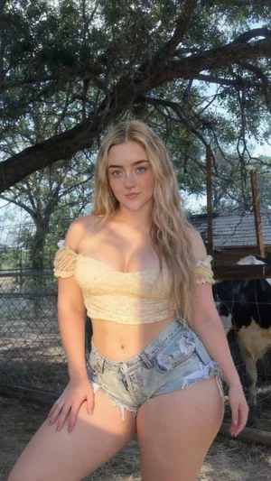 Woman with long blonde hair in off-shoulder top and denim shorts, posing outdoors near a tree and