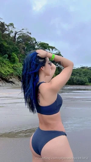 Woman with blue hair in navy bikini standing on beach, back view, tropical setting.