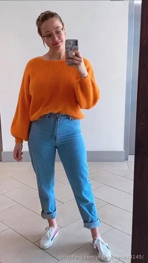 Woman in orange sweater and blue jeans takes mirror selfie indoors.