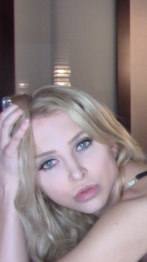 Blonde woman with blue eyes posing indoors with hand in hair.