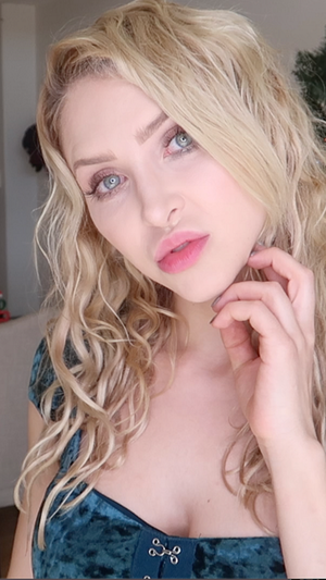 Blonde woman with blue eyes in teal velvet top indoors.