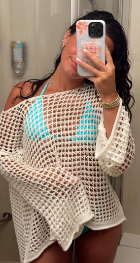 Woman in blue bikini under white crocheted coverup, taking mirror selfie in bathroom.