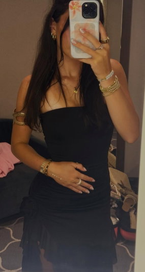 Woman in black strapless dress with jewelry, taking mirror selfie in bedroom.