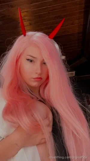 Woman with pink wig and red horns, wearing white top, posing indoors.