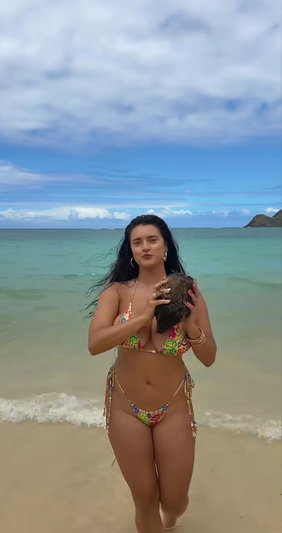 Woman in colorful bikini holding coconut on sandy beach near ocean.