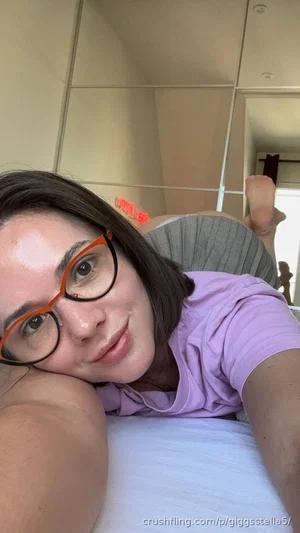Woman with glasses lying on bed, wearing purple shirt and gray bottoms, indoor setting.