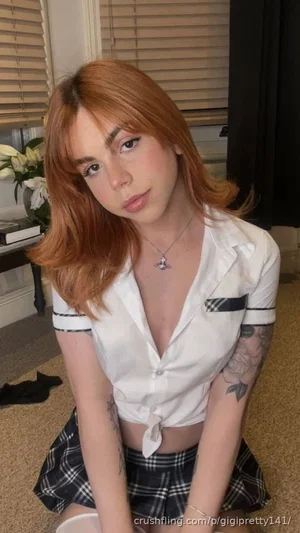 Woman with red hair in a white shirt and plaid skirt, sitting indoors with tattoos visible on arms.