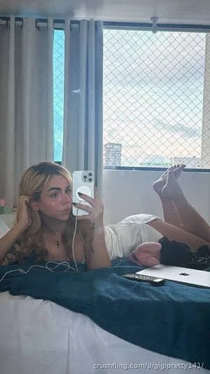 Woman with blonde hair lying on bed, wearing white top, holding phone for mirror selfie.