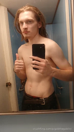 Shirtless man with long hair taking mirror selfie in bathroom, wearing dark pants and belt.