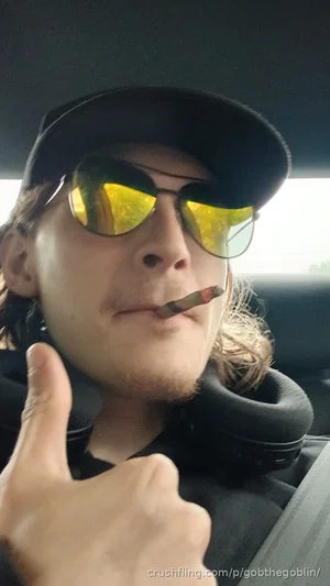 Man with long hair and sunglasses in car, giving thumbs up, wearing cap and neck pillow.