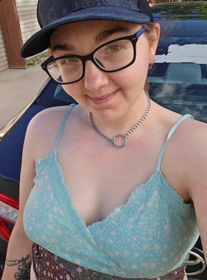 Woman in glasses and cap wearing blue lace top near car.