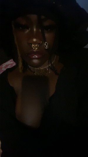 Woman with dark skin, gold jewelry, black top, and hood in dim lighting.