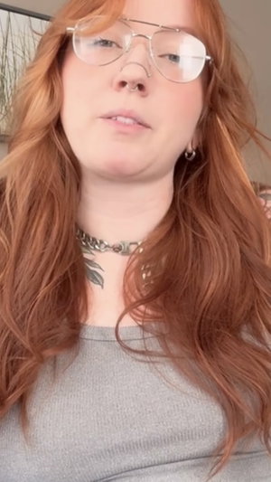 Redhead woman in glasses, choker, gray top indoors.