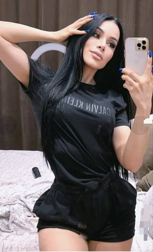 Woman with long black hair in black t-shirt and shorts taking mirror selfie in bedroom.