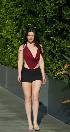 Woman in red sequined top and black shorts walking outdoors near green hedge.