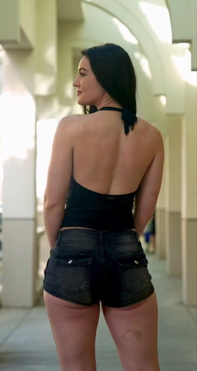 Woman with dark hair in black backless top and shorts, standing in a sunlit outdoor corridor.