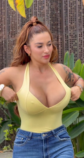 Woman in yellow top and jeans, standing outdoors with plants behind her.